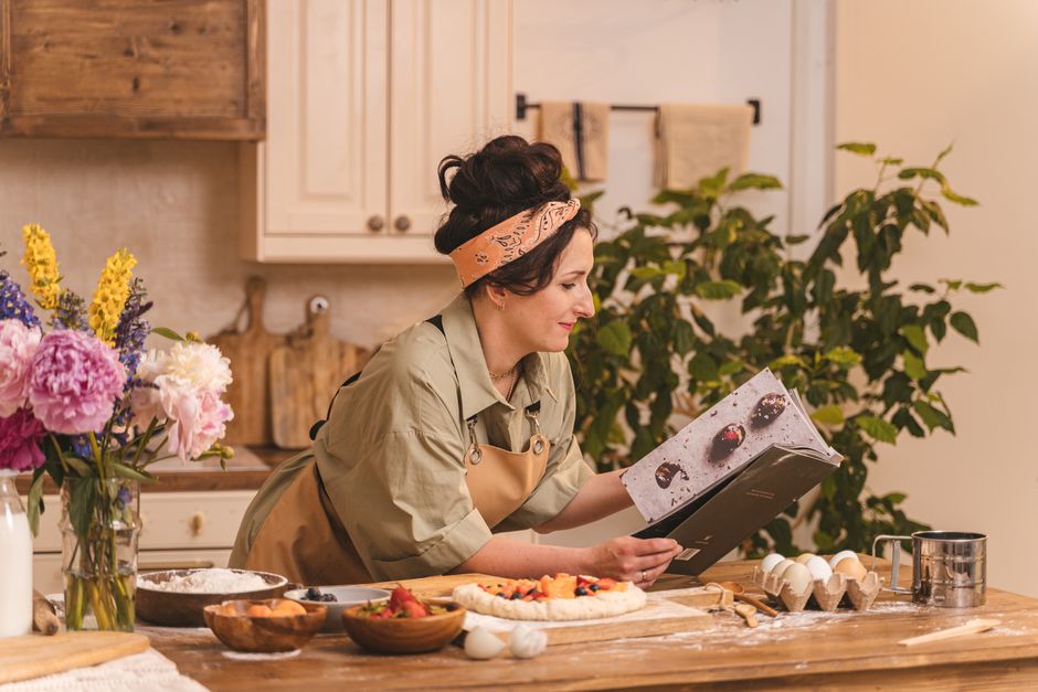 A woman looks at a cookbook while preparing pastry in a cozy kitchen setting with flowers nearby.